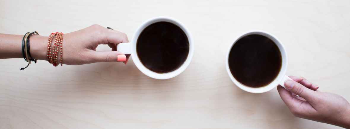 seen from above, two cups of coffee on a table, with hands of two different women visible holding the mug handles