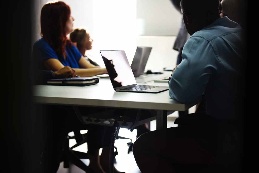 Some people sitting at a desk with laptops and notebooks watching someone (unseen) at the front of the room